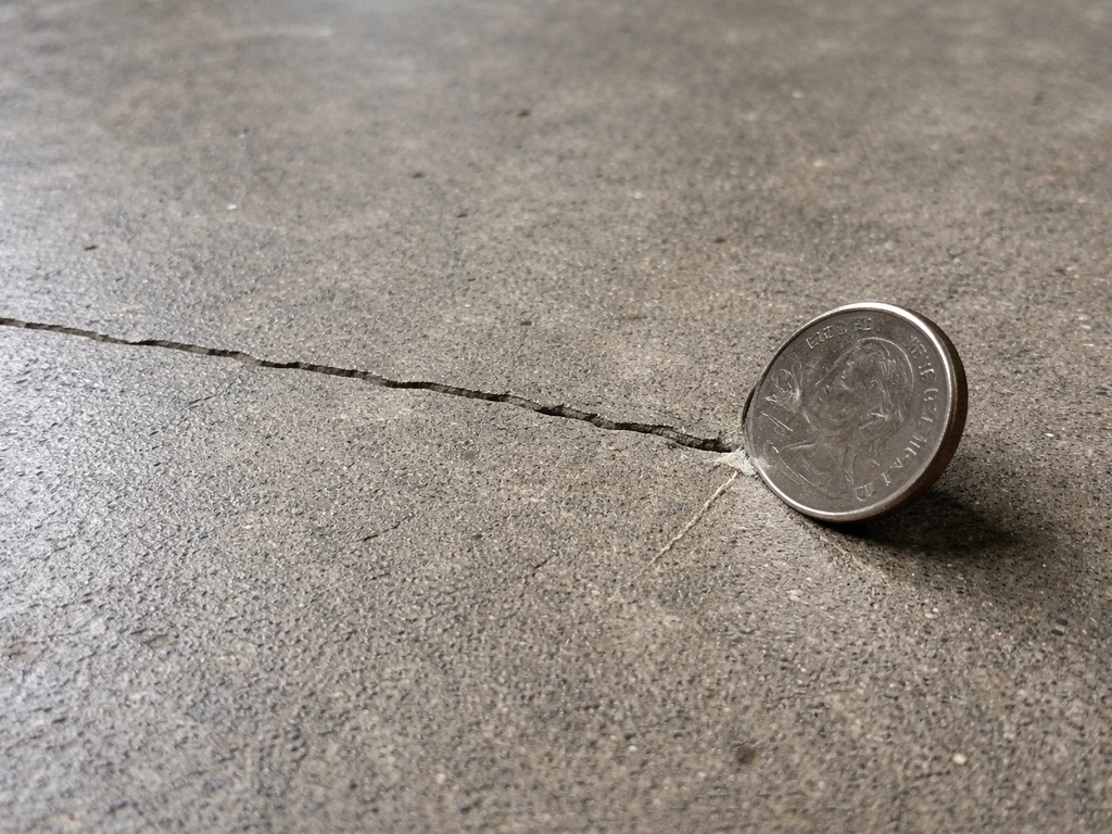 Close-up of a coin scratch test near a small hairline crack in rough concrete flooring